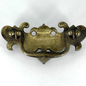 May include: A brass drawer pull with an ornate design. The pull has a curved handle and two decorative loops on each side.