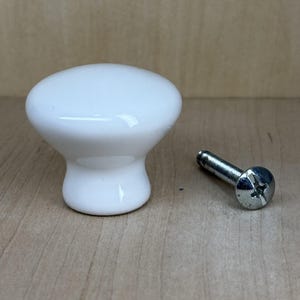 May include: A white ceramic cabinet knob with a rounded top and a flared base. A silver screw is positioned next to the knob. The knob is likely for a drawer or cabinet.