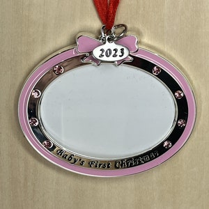 May include: Pink and silver oval-shaped photo frame ornament with a pink bow and the year 2023 on it. The frame has a white center for a photo and the words "Baby's First Christmas" around the bottom.