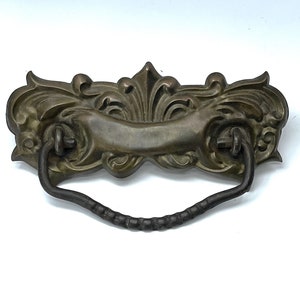 May include: Ornate brass drawer pull with a decorative scroll design and a black metal handle.