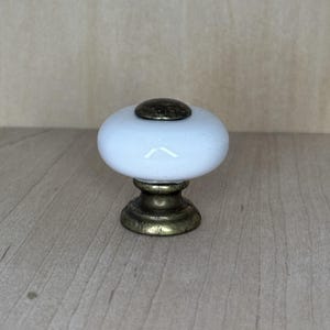 May include: A white ceramic cabinet knob with a bronze-colored base and top. The knob has a rounded, oval shape and is designed for use on drawers or cabinets. The hardware is a classic design and is suitable for various decor styles.
