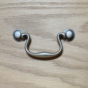 May include: A silver-colored drawer pull with a curved handle and circular mounting plates. The hardware is set against a light-colored wood grain surface, providing a rustic aesthetic.