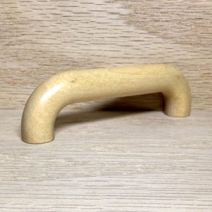 May include: A light-colored wooden cabinet handle with a curved design. The handle is made of a light-colored wood and has a smooth, rounded shape. It is designed to be mounted on a cabinet door or drawer.