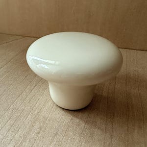 May include: A white ceramic cabinet knob with a rounded top and a cylindrical base. The knob has a glossy finish and is designed for use on drawers or cabinets. The knob is a neutral color and can be used in a variety of settings.