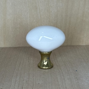 May include: A white oval cabinet knob with a gold-colored base. The knob has a smooth, glossy finish and is designed for use on drawers or cabinets. The gold base has a slightly aged appearance.