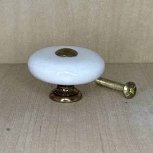 May include: White ceramic cabinet knob with a brass base and a brass screw. The knob has a rounded, oval shape with a small brass accent on top. The screw is gold-colored.