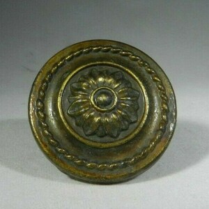 May include: A round, brass-colored door knob with a decorative floral design. The knob has a rope-like border around the edge.
