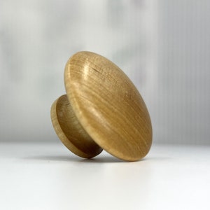 May include: A light brown wooden knob with a round top and a smaller round base. The knob is made of wood with a natural grain pattern.