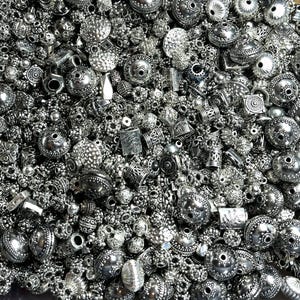 May include: A large assortment of silver metal beads and charms. The beads are of various shapes and sizes, including round, square, and oval. Some of the beads have intricate designs, while others are plain.