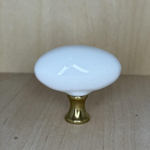 May include: White oval cabinet knob with a brass base. The knob has a smooth, glossy finish. This decorative hardware is suitable for drawers, cabinets, and furniture. The knob is approximately 2 inches long.