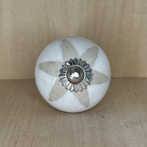 May include: A white ceramic cabinet knob with a starburst design in light beige. The knob has a silver metal base and is set against a light brown wooden surface. This decorative hardware is suitable for drawers and cabinets.