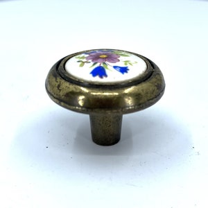 May include: A brass cabinet knob with a white ceramic top featuring a floral design in shades of purple, blue, and yellow.