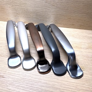 May include: Five cabinet pulls in various metallic finishes, including silver, bronze, and dark gray. The pulls have a curved, ergonomic design and are arranged on a light wood surface. These hardware pieces are suitable for kitchen or bathroom cabinets.