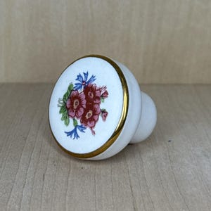 May include: White ceramic cabinet knob with a floral design. The knob features a gold rim and a cluster of red, blue, and green flowers. The knob is a decorative hardware element for furniture.