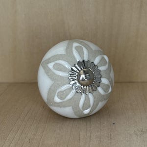 May include: A white and gray ceramic cabinet knob with a floral design. The knob has a silver-colored metal base and a rounded shape. The floral pattern is in a lighter gray color, and the knob is on a wooden surface.