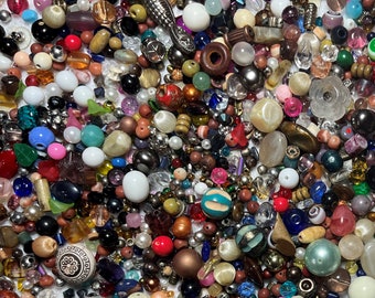 May include: A collection of various beads in different shapes, sizes, and colors. The beads include round, oval, and irregular shapes, with colors ranging from red, blue, and green to white, black, and gold. Some beads have a shiny, reflective surface, while others are matte.