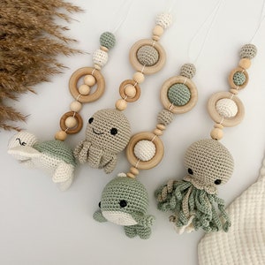 Könnte beinhalten: Drei Holz-Beißringe mit gehäkelten Tierfiguren. Die Beißringe sind in Grün und Weiß gehalten und beinhalten eine Schildkröte, einen Oktopus und einen Wal.