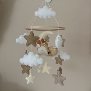 Op de afbeelding: Een houten babymobiel met een hert, wolken en sterren. Het hert is bruin en heeft een wit gezicht. De wolken zijn wit en de sterren zijn bruin en beige.