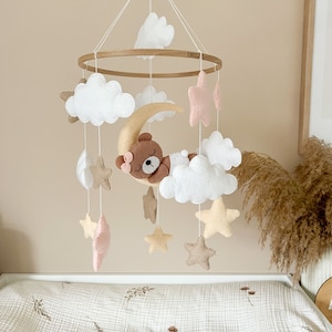 Pode incluir: Um móbile de bebé de madeira com um urso castanho a dormir numa lua crescente, nuvens brancas e estrelas bege e cor-de-rosa. O móbile está pendurado num teto branco.
