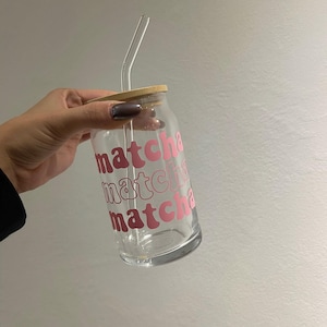 May include: A clear glass bottle with a wooden lid and a pink "matcha" graphic. A clear glass straw is inserted through the lid.