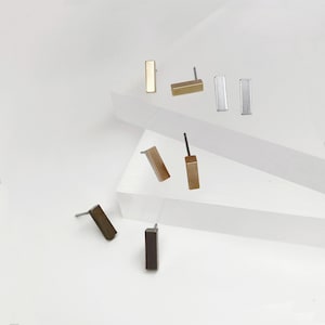 May include: A selection of rectangular stud earrings in gold, silver, and bronze finishes. The earrings are presented on a white surface, highlighting their minimalist design and geometric shapes. The studs are small and suitable for everyday wear, with a simple, modern aesthetic.