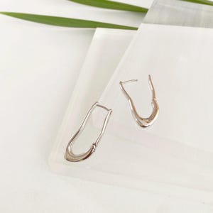 Può includere: Un paio di orecchini a cerchio argentati di forma astratta. Gli orecchini hanno una finitura liscia e lucida e un design unico e fluido. Sono esposti su una superficie bianca con una foglia verde sullo sfondo. Un accessorio di gioielleria moderno.