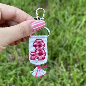 Op de afbeelding: Een rechthoekige geweven sleutelhanger met een witte achtergrond en een roze en donker magenta gepixeld dinosaurusmotief. Het textielstuk is bevestigd aan een zilverkleurige metalen karabijnhaak en afgewerkt met een kleine witte en roze kwast.