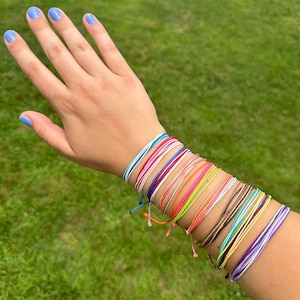 Könnte beinhalten: Eine Sammlung farbenfroher Schnurarmbänder in verschiedenen Farbtönen, darunter Blau, Rot, Rosa, Orange, Grün und Lila. Die Armbänder werden an einem Handgelenk vor dem Hintergrund von grünem Gras getragen. Jedes Armband besteht aus dünnen, gewebten Fäden.