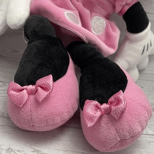Puede incluir: Primer plano de un par de zapatillas de felpa rosa con la parte superior negra y lazos rosas con lunares blancos. Las zapatillas forman parte de un disfraz m&aacute;s grande, con un atuendo rosa y guantes blancos visibles en el fondo.