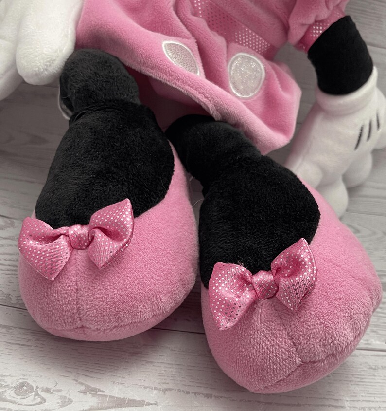 Puede incluir: Primer plano de un par de zapatillas de felpa rosa con la parte superior negra y lazos rosas con lunares blancos. Las zapatillas forman parte de un disfraz m&aacute;s grande, con un atuendo rosa y guantes blancos visibles en el fondo.