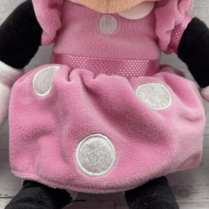Puede incluir: Mu&ntilde;eca de peluche de Minnie Mouse. La mu&ntilde;eca lleva un vestido rosa con lunares blancos y un lazo rosa a juego. El vestido tiene un cintur&oacute;n rosa brillante. La mu&ntilde;eca tiene brazos y piernas negros y guantes blancos.