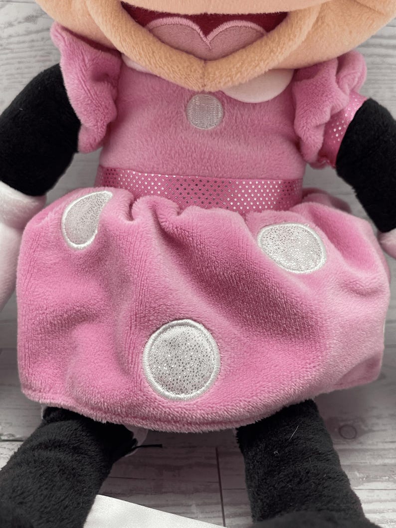 Puede incluir: Mu&ntilde;eca de peluche de Minnie Mouse. La mu&ntilde;eca lleva un vestido rosa con lunares blancos y un lazo rosa a juego. El vestido tiene un cintur&oacute;n rosa brillante. La mu&ntilde;eca tiene brazos y piernas negros y guantes blancos.