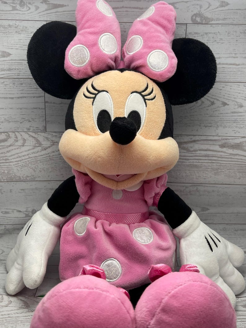 Puede incluir: Un mu&ntilde;eco de peluche de Minnie Mouse. El mu&ntilde;eco tiene un vestido y un lazo rosas con lunares blancos, orejas negras y guantes blancos. El mu&ntilde;eco tiene una cara bronceada con ojos negros y una nariz negra. El mu&ntilde;eco es un juguete suave y tierno.