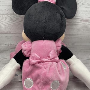 Puede incluir: Una mu&ntilde;eca de peluche de Minnie Mouse, vista desde atr&aacute;s, con cabeza y orejas negras, un vestido rosa con lunares blancos y un lazo a juego. La mu&ntilde;eca tiene guantes blancos y una cola negra.