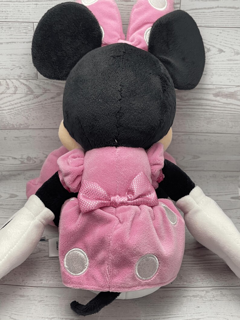 Puede incluir: Una mu&ntilde;eca de peluche de Minnie Mouse, vista desde atr&aacute;s, con cabeza y orejas negras, un vestido rosa con lunares blancos y un lazo a juego. La mu&ntilde;eca tiene guantes blancos y una cola negra.