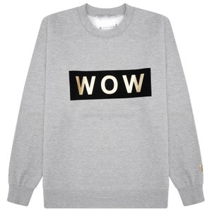 Puede incluir: Una sudadera gris claro con la palabra "WOW" en letras doradas sobre un rectángulo negro en el pecho. La sudadera tiene cuello redondo y mangas largas. El logotipo de la marca es visible en la parte superior de la espalda y el puño.