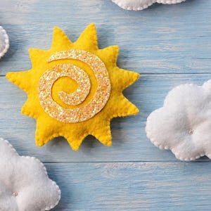 Può includere: Un sole in feltro giallo con una spirale centrale di glitter dorati e tre nuvole in feltro bianco con piccoli accenti argentati. Il sole presenta raggi appuntiti e una consistenza morbida, mentre le nuvole hanno un aspetto trapuntato.