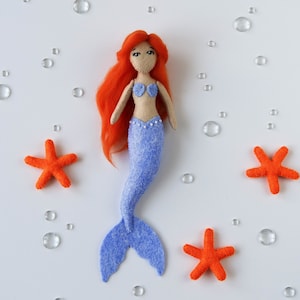 May include: A handmade felt mermaid doll with long red hair, a blue tail, and a seashell bra. The doll is surrounded by orange felt starfish and clear plastic bubbles.