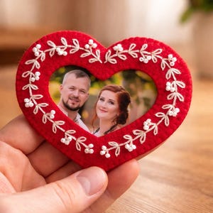 May include: A red felt heart-shaped photo frame featuring white embroidered vine patterns and small white beads. The center contains a photograph of a man and a woman. The frame is constructed from soft felt material with detailed stitching.