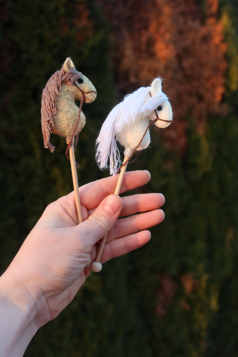 Puede incluir: Dos caballos de palo de madera, uno marr&oacute;n y otro blanco, se sostienen en la mano de una persona. Los caballos est&aacute;n hechos de fieltro y tienen una crin y una cola.