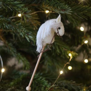 Puede incluir: Adorno de caballo de balanc&iacute;n de fieltro blanco colgado de un &aacute;rbol de Navidad con luces blancas.