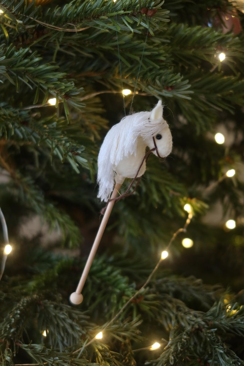 Puede incluir: Adorno de caballo de balanc&iacute;n de fieltro blanco colgado de un &aacute;rbol de Navidad con luces blancas.