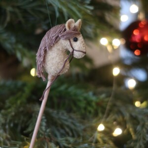 Puede incluir: Un adorno de cabeza de caballo de fieltro marr&oacute;n y blanco colgado de un &aacute;rbol de Navidad con luces blancas. El caballo tiene una crin y una cola marrones y una brida de cuero marr&oacute;n.