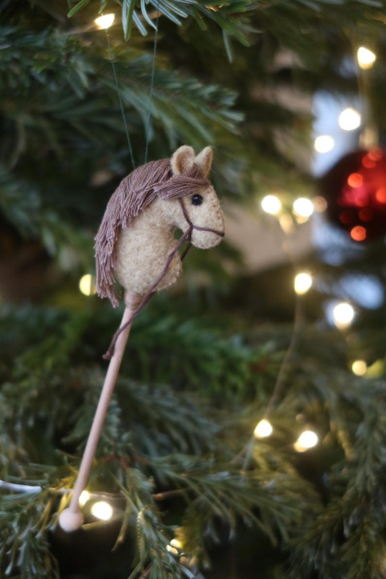 Puede incluir: Un adorno de cabeza de caballo de fieltro marr&oacute;n y blanco colgado de un &aacute;rbol de Navidad con luces blancas. El caballo tiene una crin y una cola marrones y una brida de cuero marr&oacute;n.