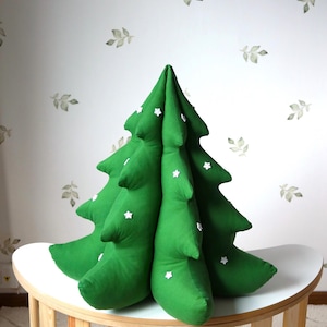 May include: A green fabric Christmas tree pillow with white snowflake embellishments. The pillow is sitting on a white table with wooden legs.