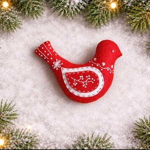 May include: A red felt bird ornament featuring white embroidered details, including a wing with a branch pattern, a dotted tail, and a small white dot for an eye. The bird is surrounded by green pine branches and white snow.