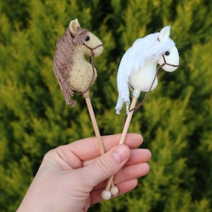 Puede incluir: Dos caballos de palo de madera en miniatura, uno marr&oacute;n y otro blanco, sostenidos en una mano.
