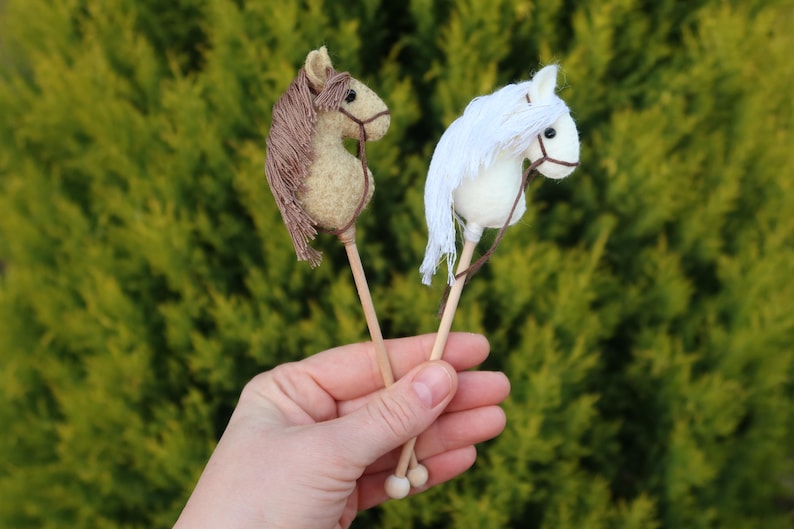 Puede incluir: Dos caballos de palo de madera en miniatura, uno marr&oacute;n y otro blanco, sostenidos en una mano.