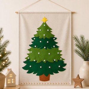 Op de afbeelding: Een rechthoekige wandhanger van beige stof met een gelaagde kerstboom van groen vilt. De boom is versierd met kleine witte knoopjes in de vorm van sneeuwvlokken en bekroond met een gele ster. Een rij kleine houten knoopjes siert de onderrand.