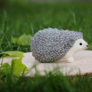 May include: A small, gray and white needle felted hedgehog figurine with a white belly and black eyes. The hedgehog is sitting on a wooden slice.